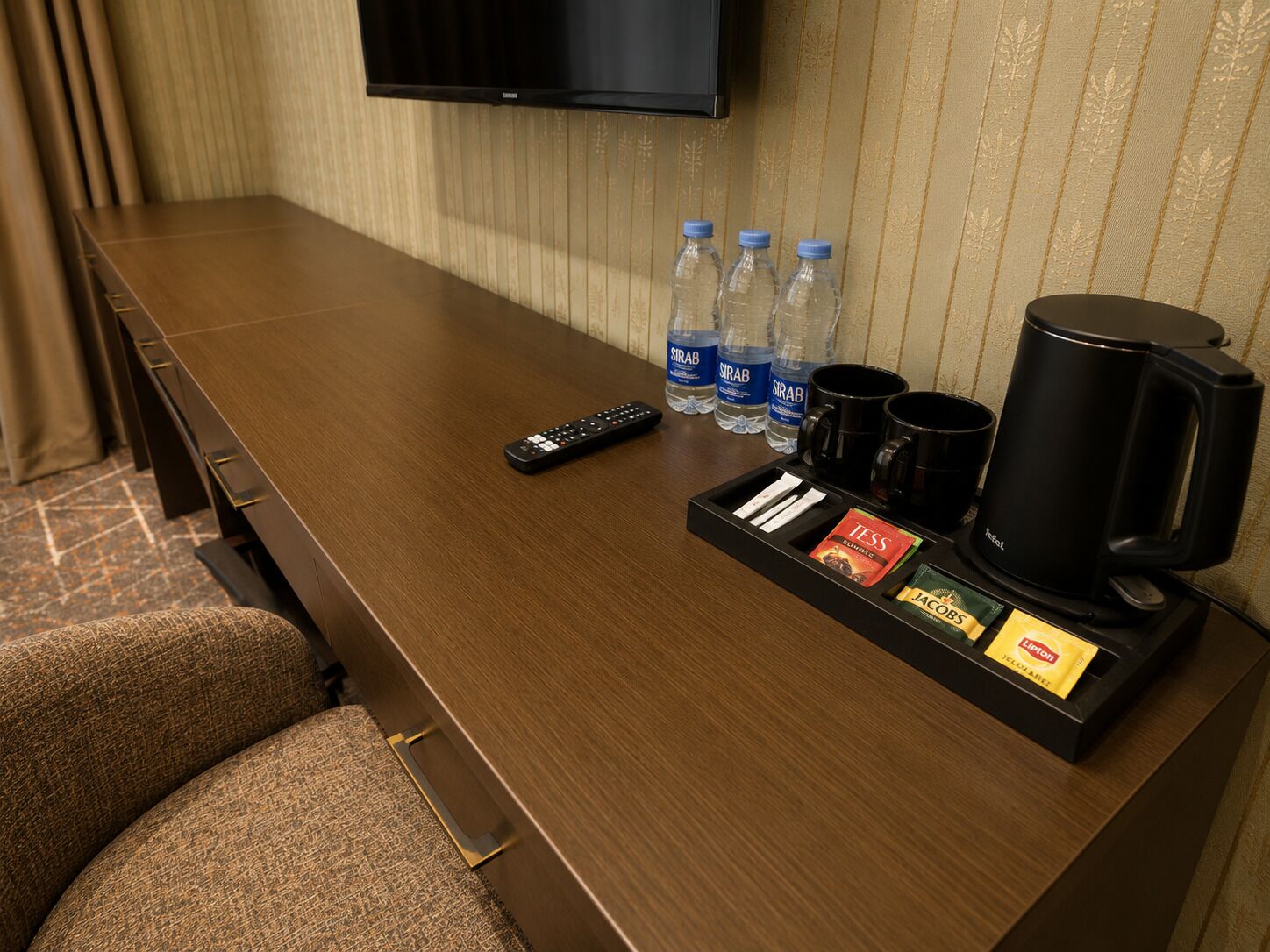 A hotel room with a wooden desk, a remote control, water bottles, coffee mugs, and a tray with snacks and a coffee maker.