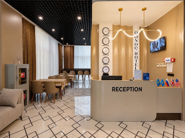 Zona de recepción de hotel con mostrador, relojes mostrando diferentes ciudades, una pantalla de televisión y exhibición de banderas.