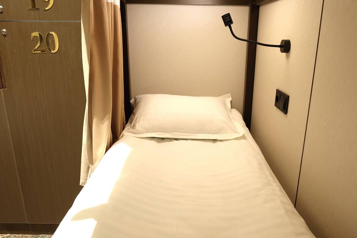 Un lit simple avec un matelas et des oreillers blancs, un luminaire fixé au mur et une porte avec une plaque numérotée.