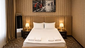 Una habitación de hotel con una cama grande, dos mesitas de noche con lámparas, un cabecero, un cuadro grande en la pared y una ventana con cortinas.