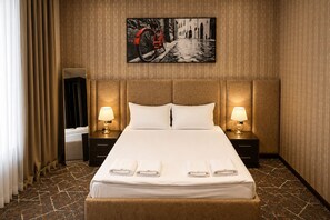 Una habitación de hotel con una cama grande, dos mesitas de noche con lámparas, un cabecero, un cuadro grande en la pared y una ventana con cortinas.