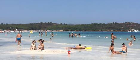 A beach with people sunbathing and swimming, clear blue water, and a hilly landscape in the background.