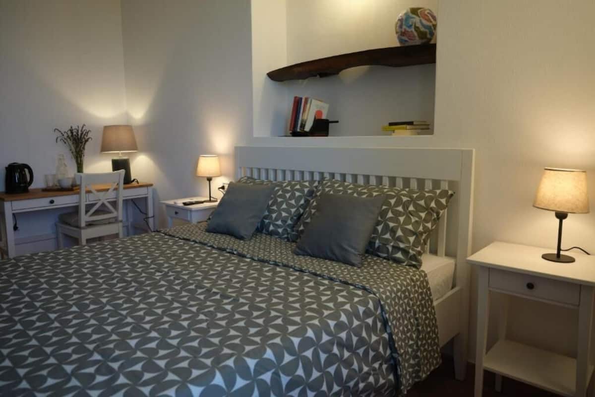 A bedroom with a bed, two bedside tables, a nightstand, a lamp, a shelf with books, and a decorative item.