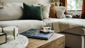 A cozy living room with a sofa, a glass of water, a book, and a wooden side table. - Alpine Ridge | Brand New Build | Sleeps 16 (Boyne City)