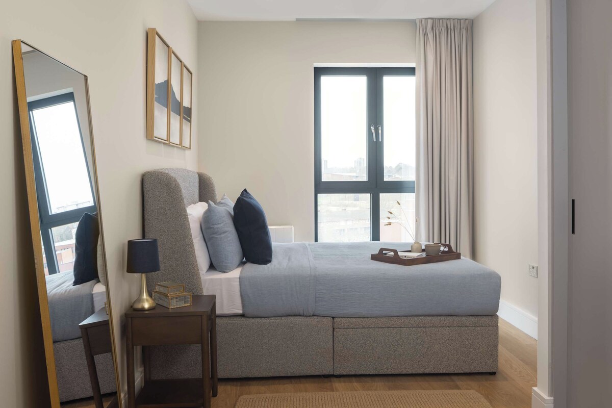A modern bedroom with a large bed, a bedside table, a mirror, and a window with curtains.
