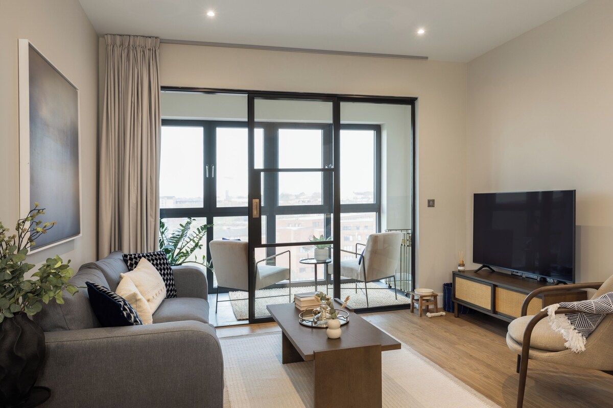 A modern living room with a sofa, armchairs, a coffee table, a TV, and a sliding glass door leading to a balcony.