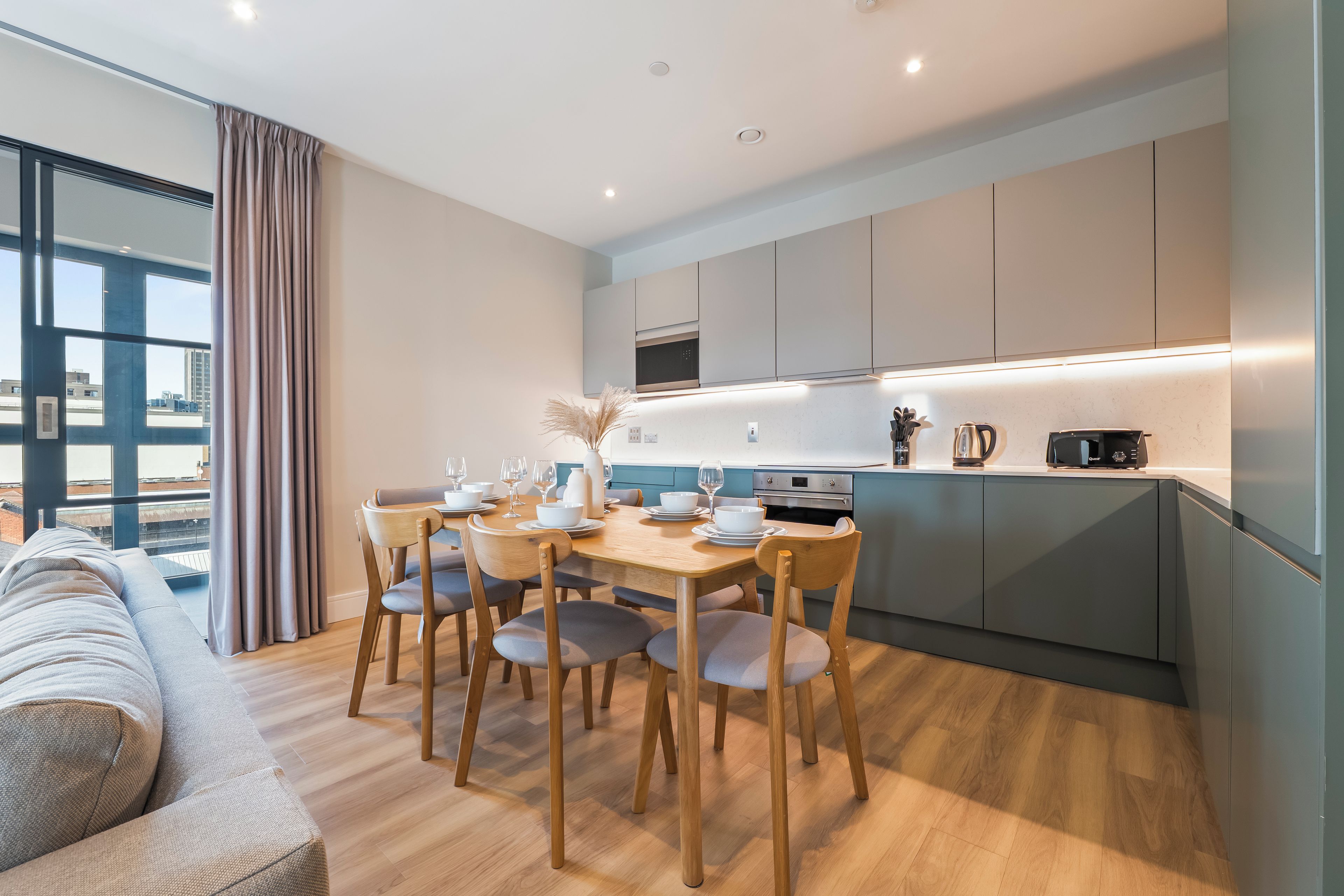 A modern kitchen with a dining area, wooden chairs, and a view through large windows.