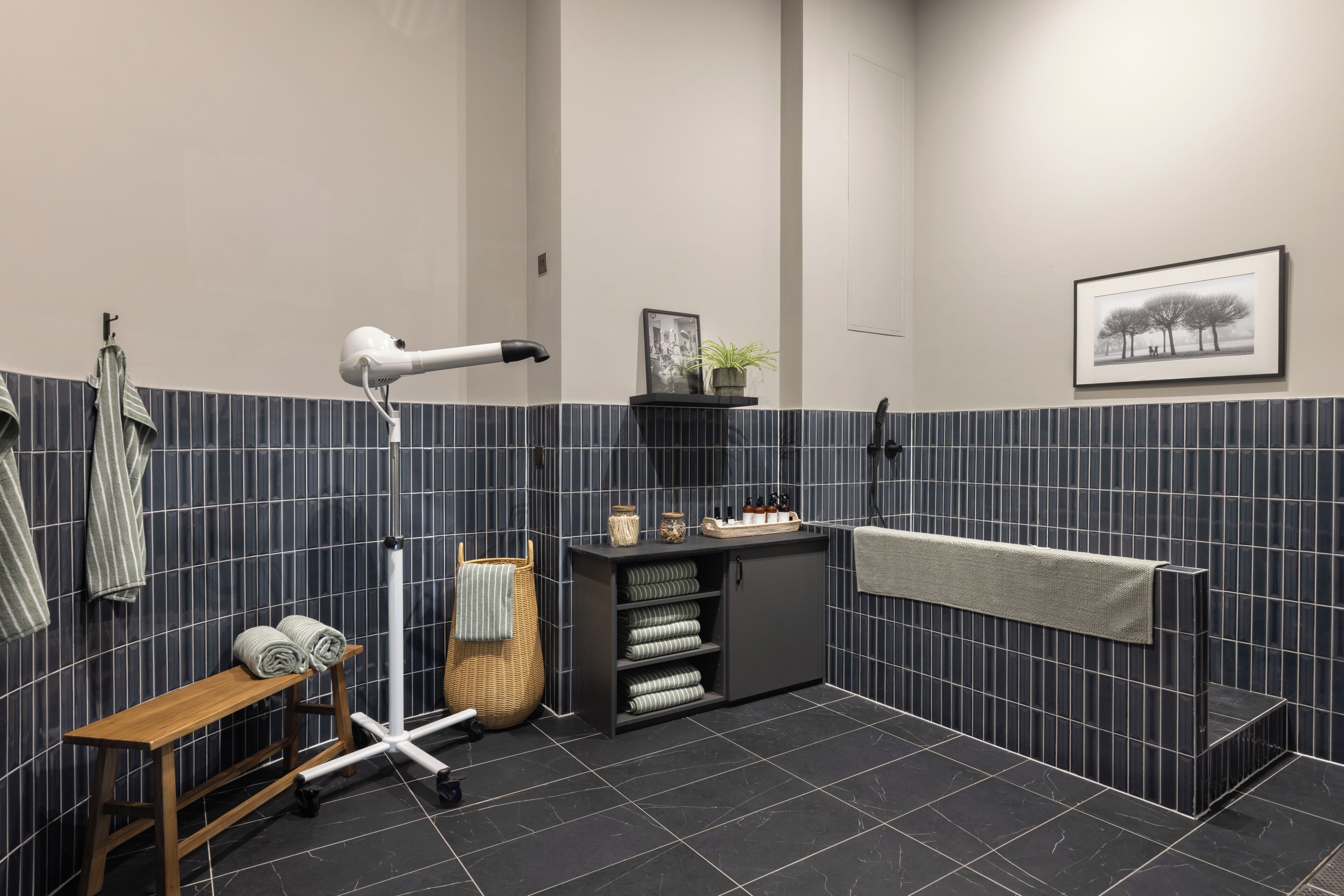 A modern bathroom with a dark tiled wall, a wooden bench, a hairdryer, a towel rack, a shelf with toiletries, and a framed picture.