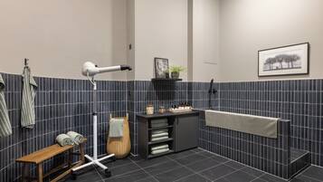 A modern bathroom with a dark tiled wall, a wooden bench, a hairdryer, a towel rack, a shelf with toiletries, and a framed picture.