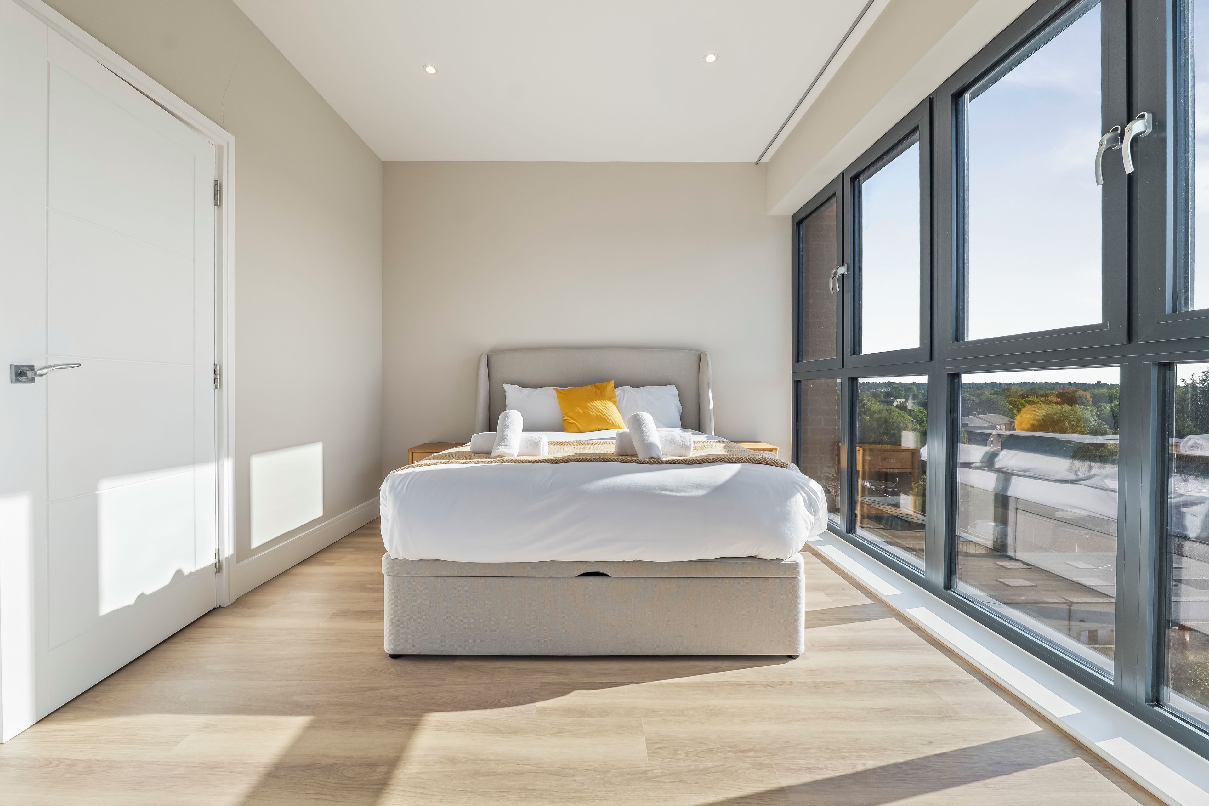 A modern bedroom with a large bed, a view through the window, and a door on the left.