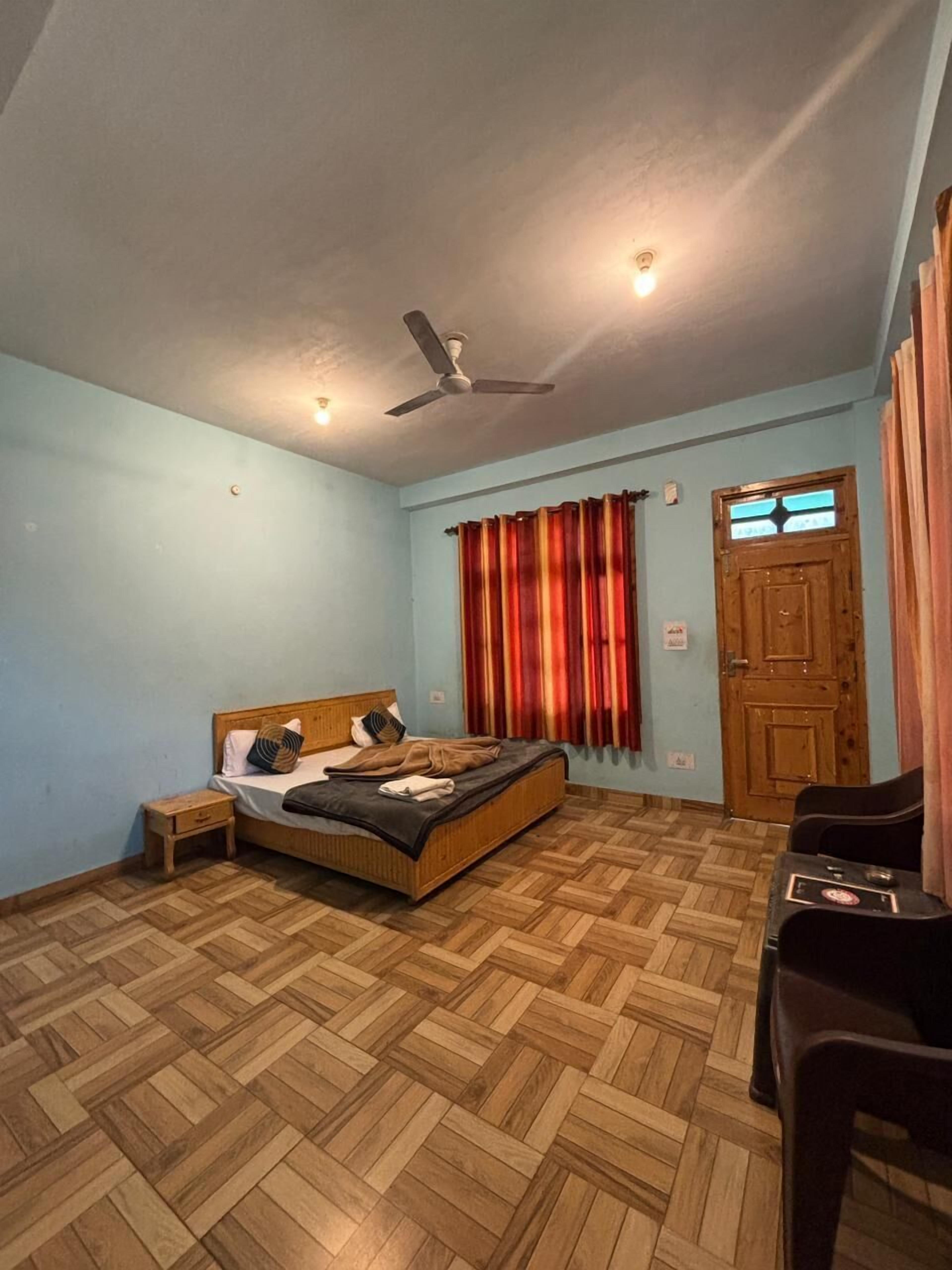 Quarto com cama, mesa de cabeceira, ventilador de teto, porta de madeira e janela com cortinas.