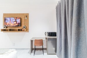 Ein modernes Zimmer mit an einer Holzvertäfelung befestigtem Fernseher, einer kleinen Küchenzeile mit Mikrowelle, einem Sessel und einem grauen Vorhang.