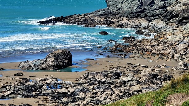 Un paisaje costero rocoso con olas rompiendo sobre la orilla, un acantilado y una playa de arena.