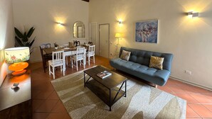 A living room with a sofa, coffee table, dining table with chairs, and a painting on the wall. - Cosy apartment for 8 guests with WIFI, A/C, TV and balcony (Lucca)