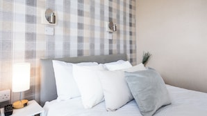 Una cama bien hecha con almohadas blancas y grises, una lámpara de noche y un control remoto sobre una mesita blanca.