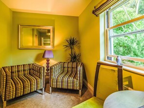Ein Zimmer mit zwei gemusterten Sesseln, einem Spiegel, einer Lampe, einer hohen Pflanze und einem Fenster mit Blick auf Grün.