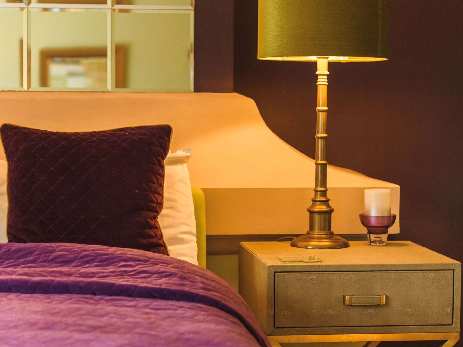 Un lit avec une couverture violette, un oreiller foncé, une table de chevet avec une lampe, une bougie et un petit tiroir.