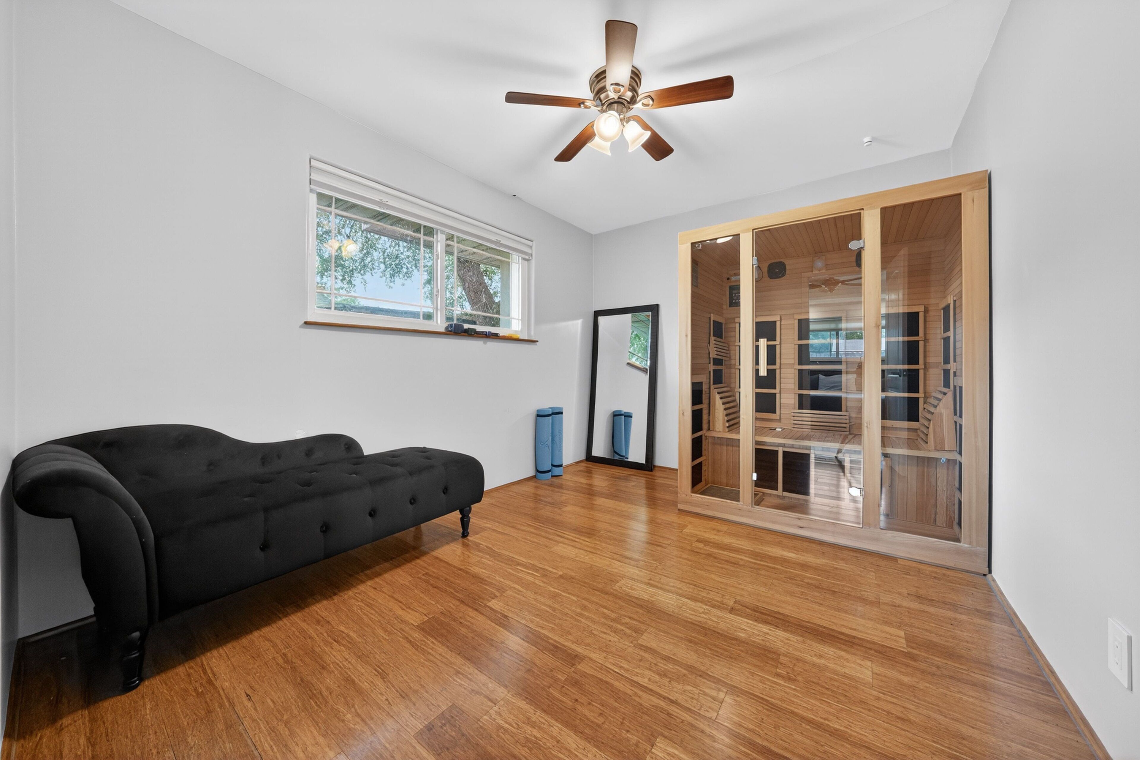 A room with a black sofa, wooden flooring, a ceiling fan, a window, and a sliding glass door leading to a wooden staircase.