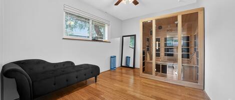 A room with a black sofa, wooden flooring, a ceiling fan, a window, and a sliding glass door leading to a wooden staircase.