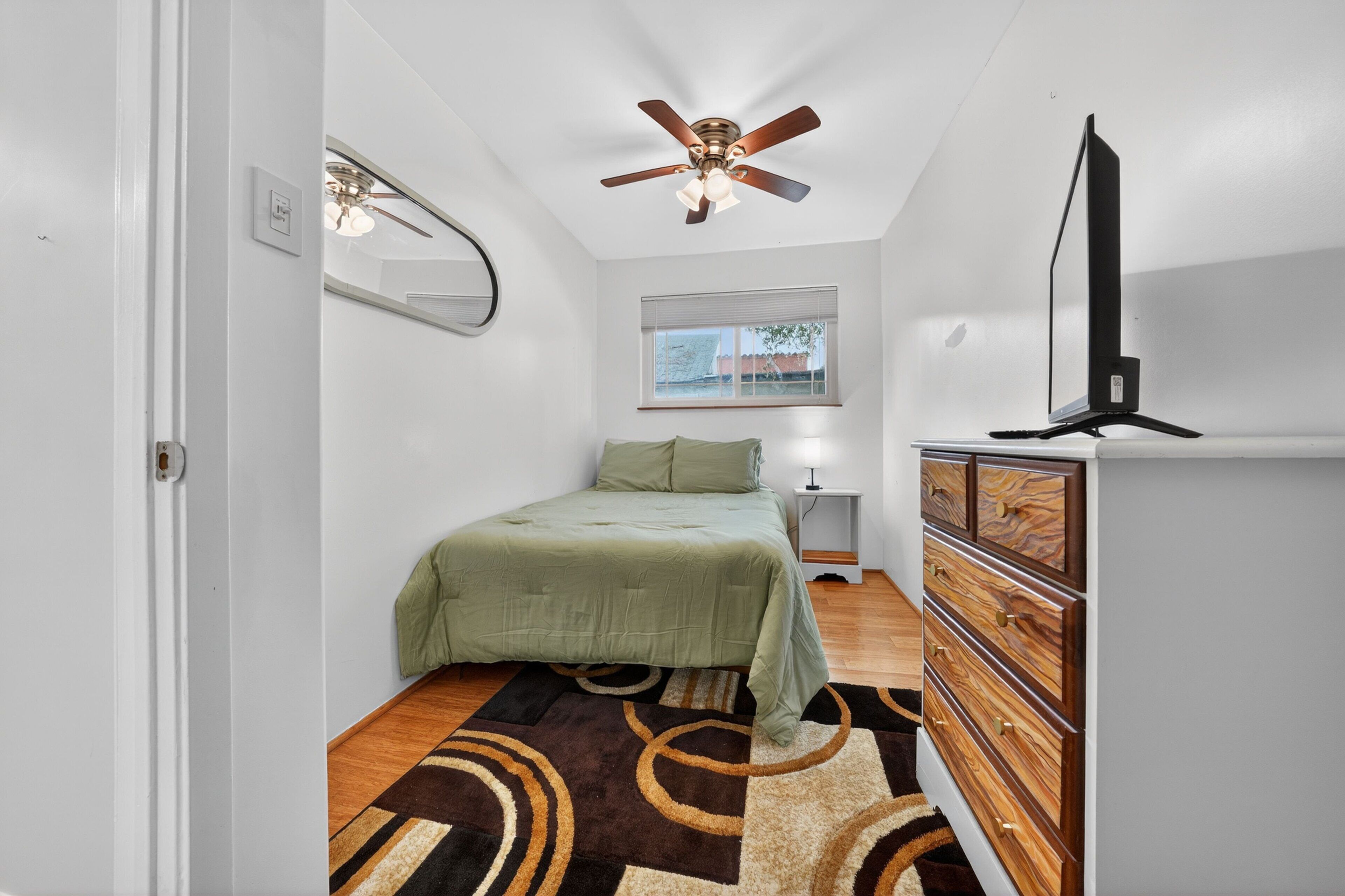 A bedroom with a bed, a ceiling fan, a window, a dresser, and a television.