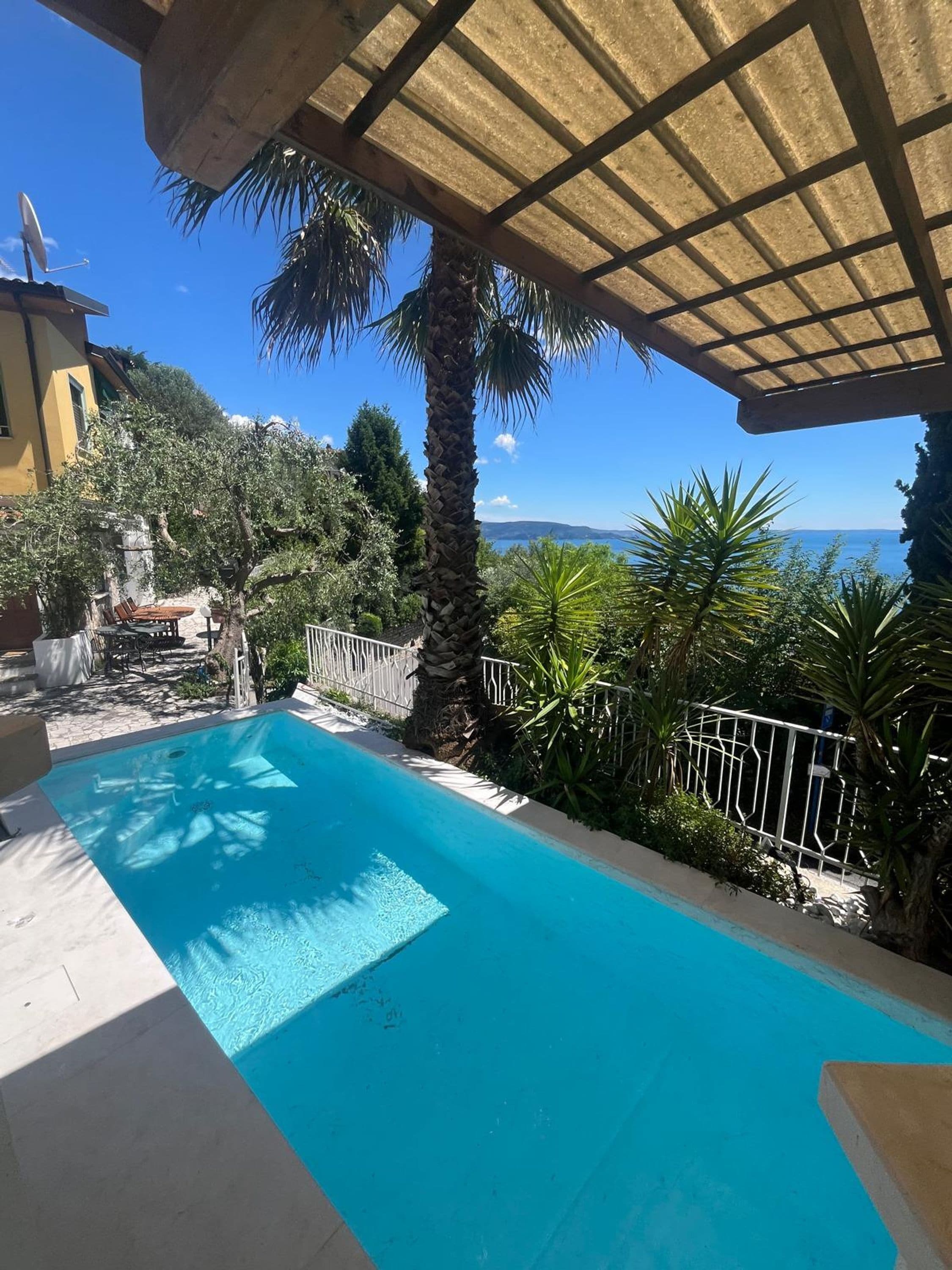 A swimming pool with a view of the sea, surrounded by greenery and a pergola.
