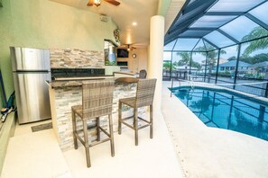 A kitchen with a stainless steel refrigerator, a stone backsplash, wicker bar stools, and a pool area with a glass enclosure. - Apollos Anchor - 926 Golf Island Dr 5 Bedroom Home (Apollo Beach)
