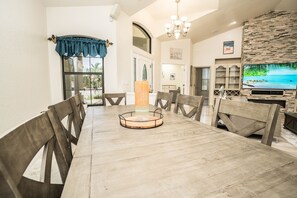 A dining area with a long wooden table, chairs, a chandelier, and a stone wall with a TV. - Apollos Anchor - 926 Golf Island Dr 5 Bedroom Home (Apollo Beach)