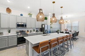 Eine moderne Küche mit weißen Schränken, Edelstahlgeräten, einer großen Kochinsel mit Barhocker und hängenden Korbtm-Pendelleuchten.