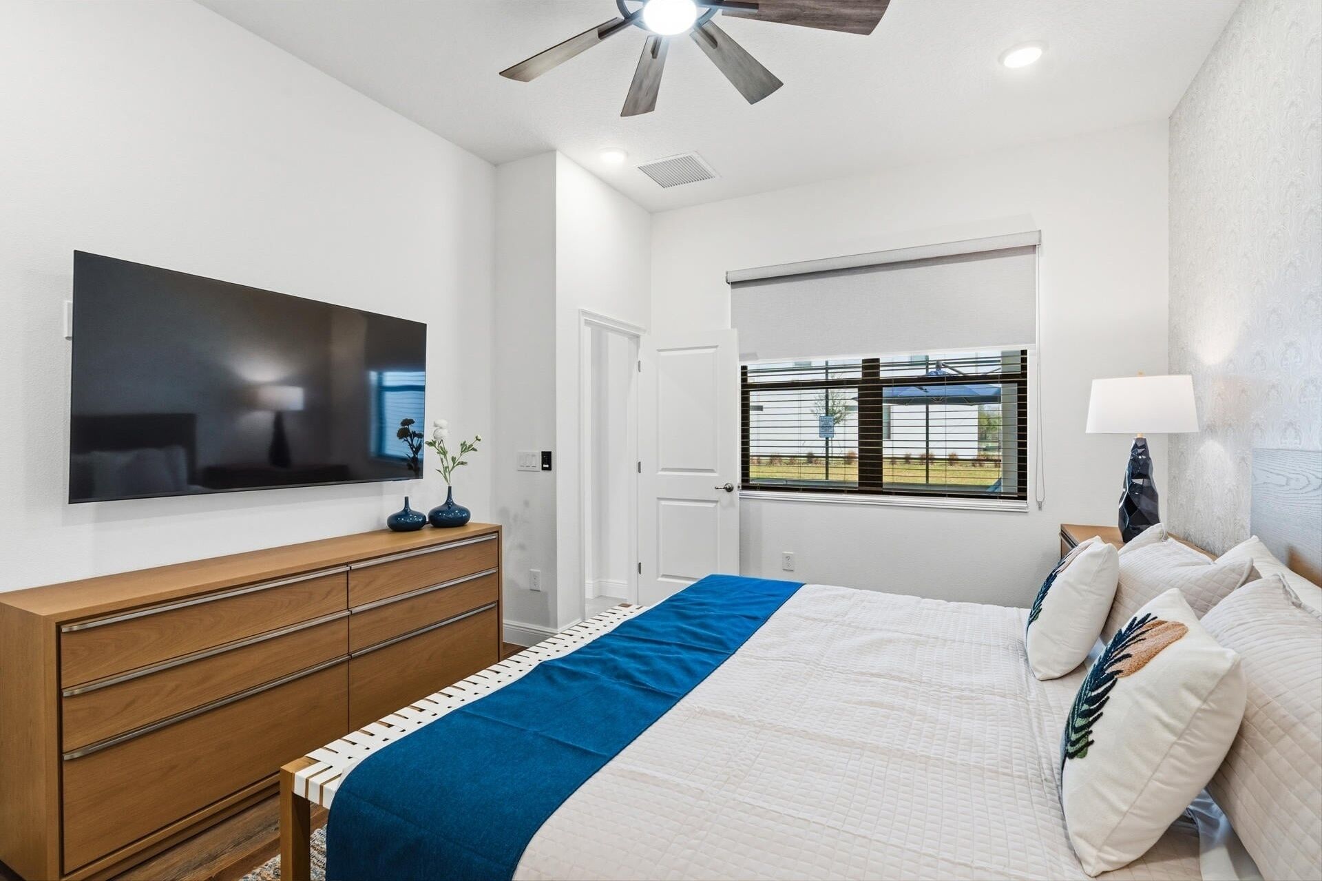 A bedroom with a bed, a dresser, a TV, a window with blinds, and a ceiling fan.