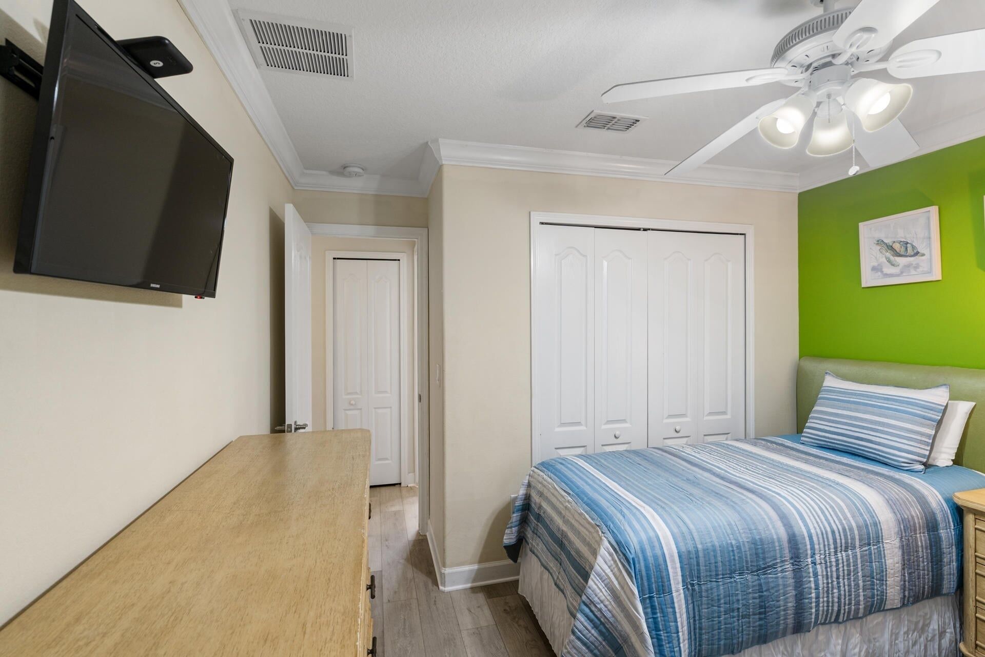 Une chambre avec un lit, un bureau, une télévision, un ventilateur de plafond et un mur vert.