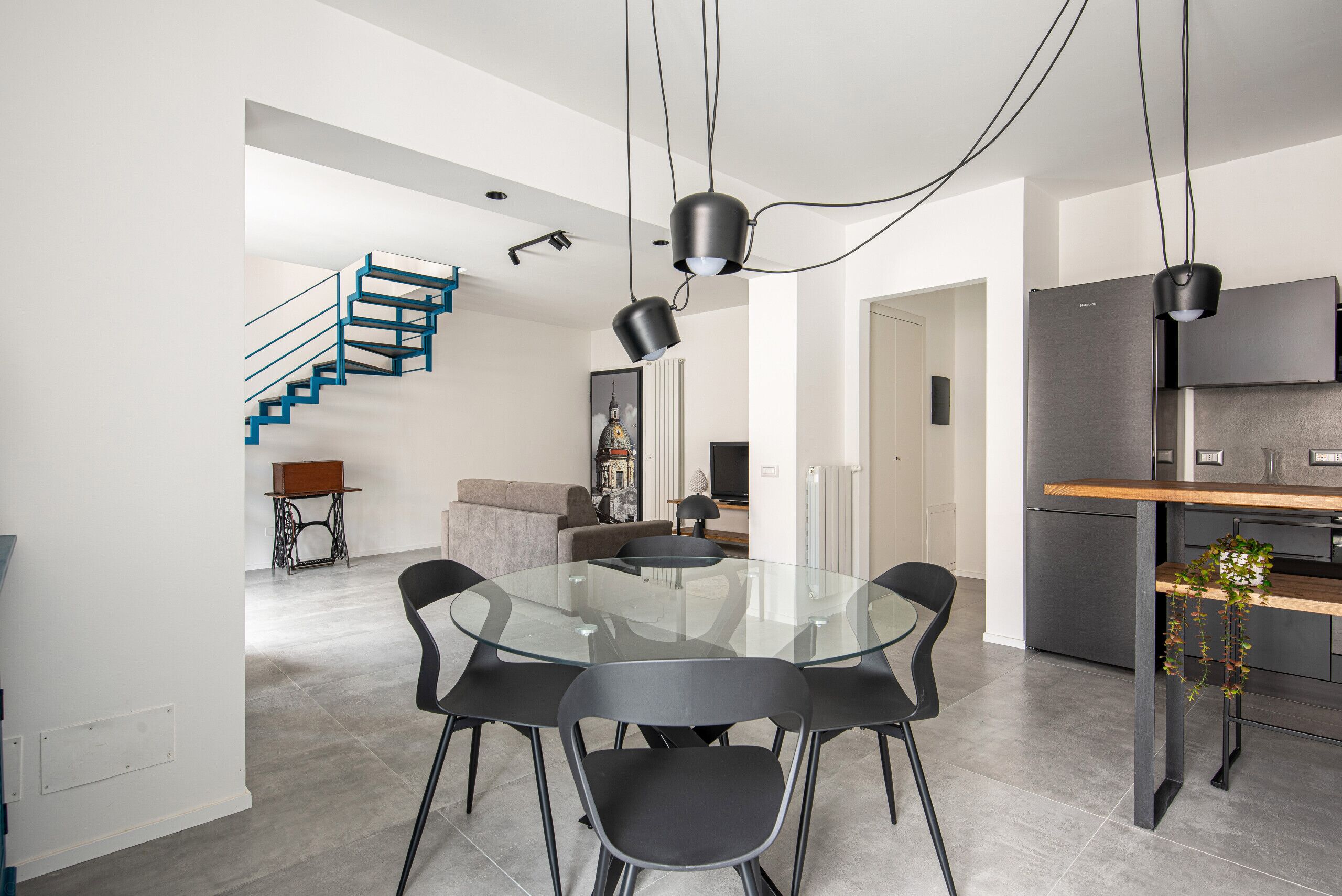 Uma cozinha moderna com geladeira embutida, um espaço de refeições com mesa e cadeiras de vidro, e uma escada com corrimão azul.