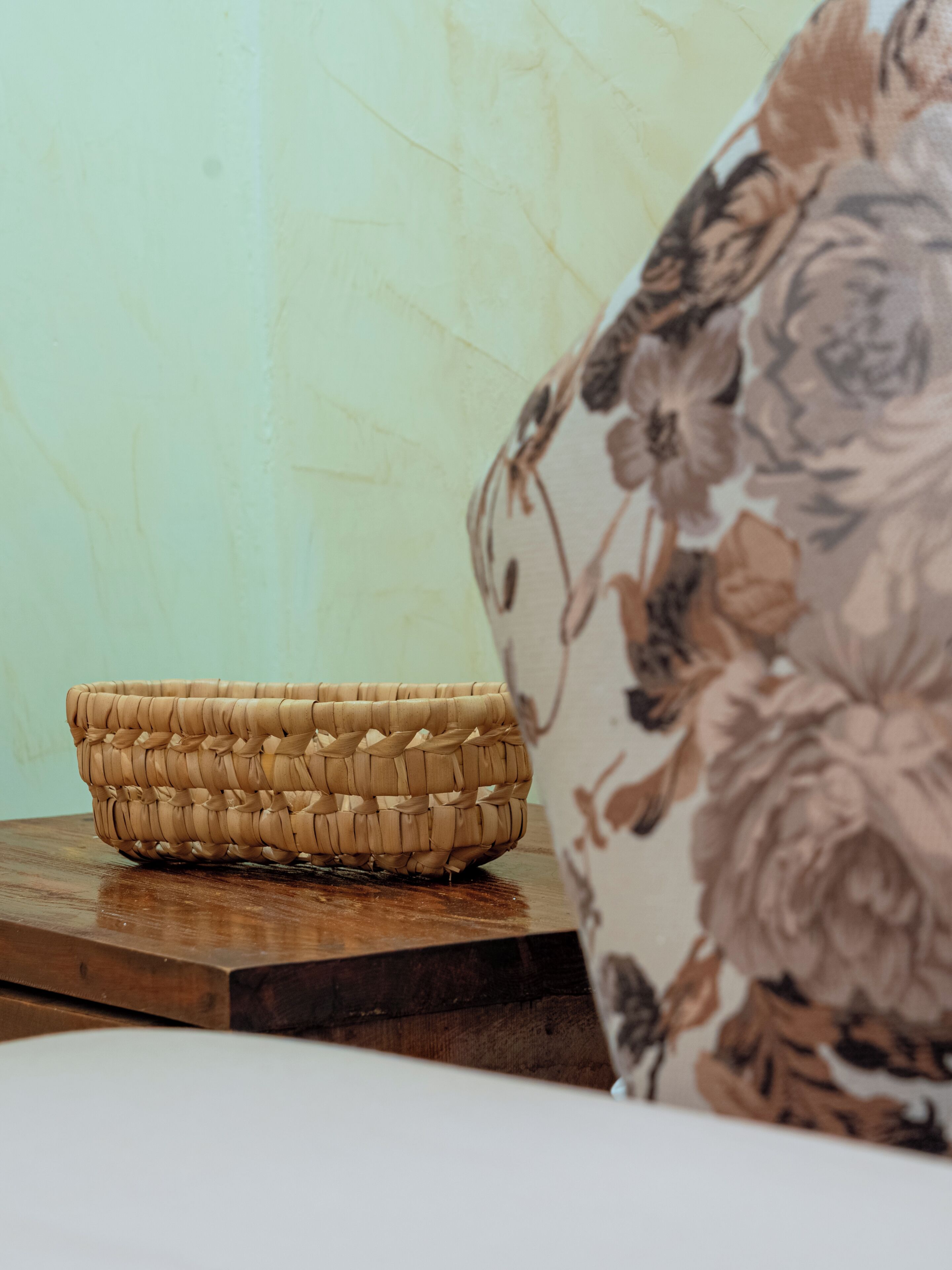 A woven basket on a wooden table with a floral-patterned cushion in the background.