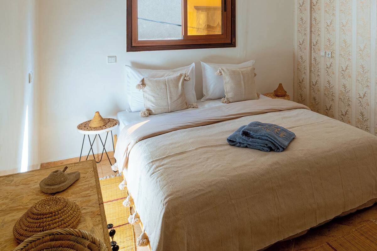 A bedroom with a bed, a window with blinds, a hanging light fixture, and a patterned wall.