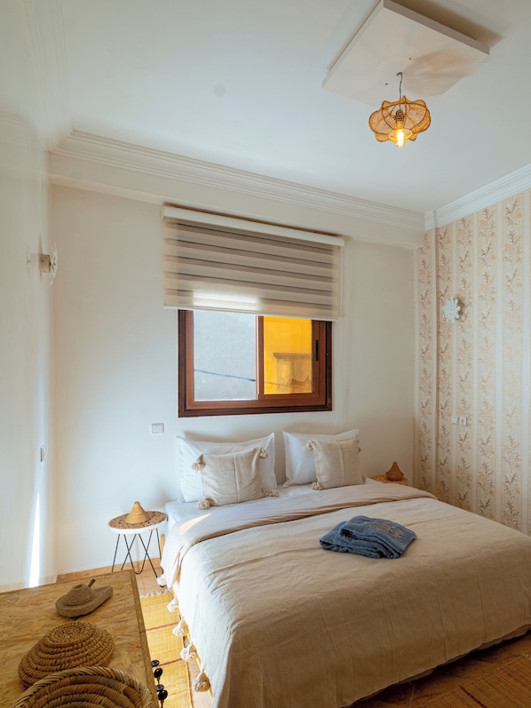 A bedroom with a bed, a window with blinds, a hanging light fixture, and a patterned wall.