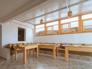 A room with a long wooden bench, a patterned floor, and large windows with a view of a hilly landscape. - Roots Surf House (Agadir)