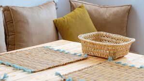 Una cesta tejida sobre una mesa de madera con individuales tejidos, acompañada de dos almohadas.