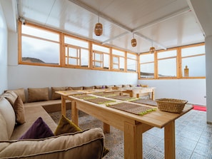 A room with a long wooden table, cushioned seating, large windows, and hanging lanterns.