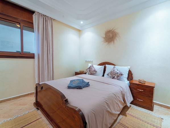 A bedroom with a wooden bed, a window with curtains, a wall-mounted light, and a decorative wall piece. - Roots Surf House (Agadir)