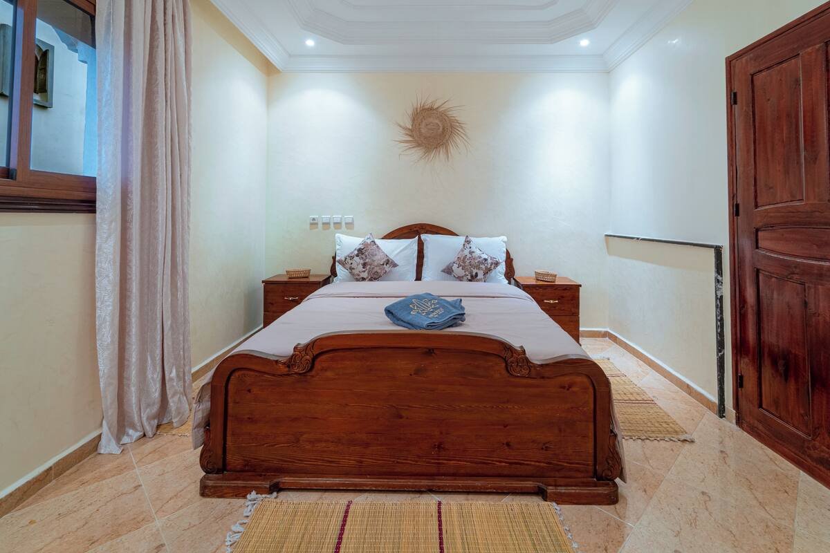 A bedroom with a wooden bed, bedside tables, a wall-mounted light fixture, and a large wooden door.