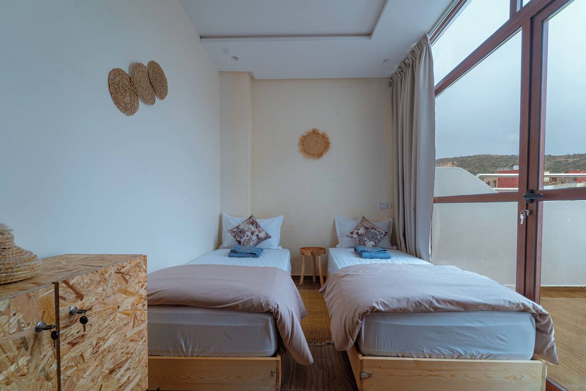 A room with two beds, a wooden cabinet, a small stool, and a large window.