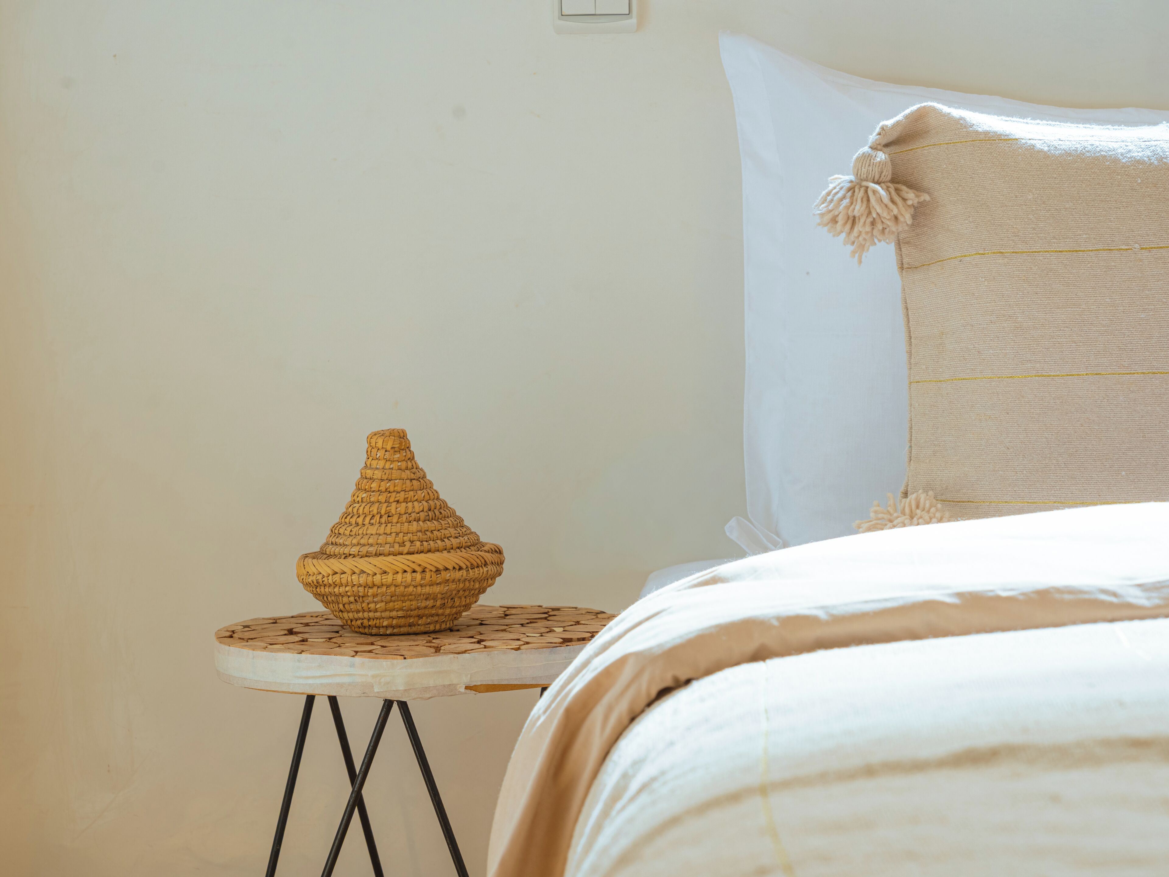 Un letto con un cuscino bianco e una coperta lavorata, un tavolino rotondo con un cesto di vimini e un interruttore a parete.