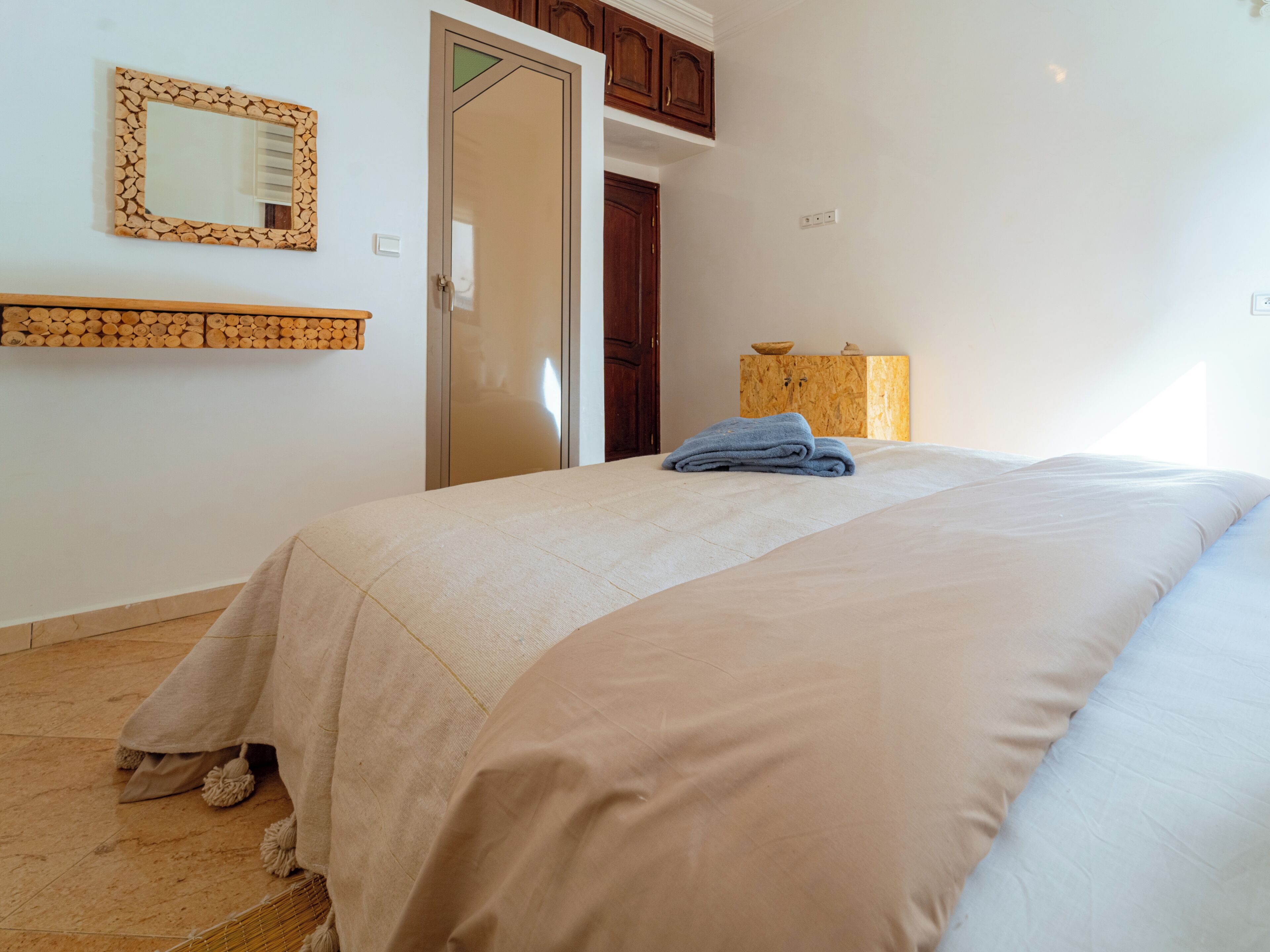 Une chambre à coucher comprenant un lit, une tête de lit en bois, un miroir, une armoire en bois et une petite table sur laquelle se trouve un bol.