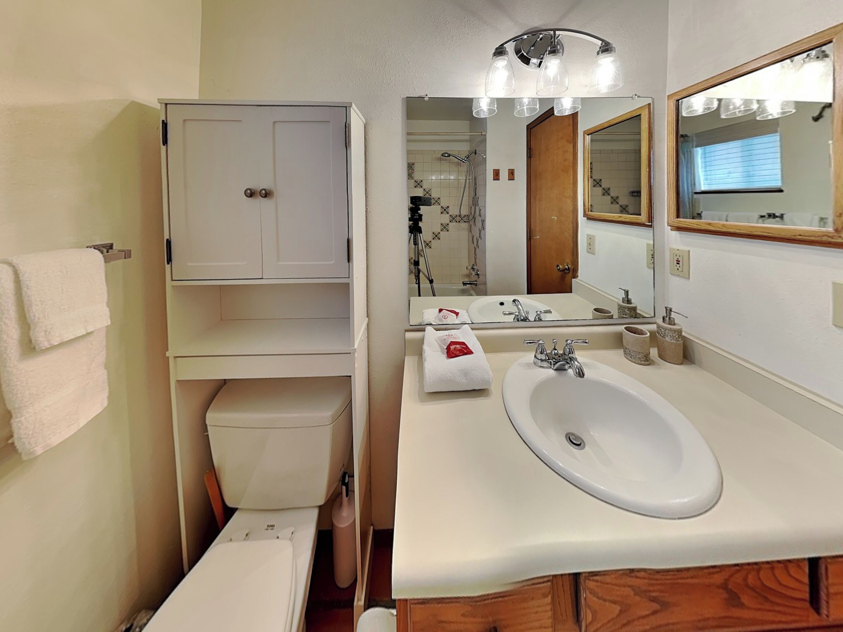 Ein Badezimmer mit weißem Waschbecken, Spiegel und Schrank. Im Hintergrund ist eine Dusche erkennbar.