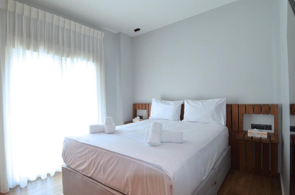 Habitación de hotel con una cama grande, cabecera de madera, mesita de noche y una ventana con cortinas translúcidas.