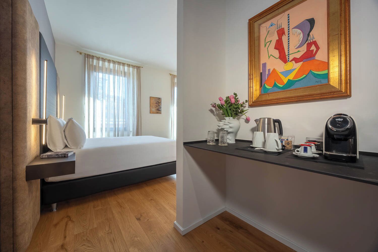 Una habitación de hotel con una cama, una cafetera, un jarrón con flores y un cuadro en la pared.