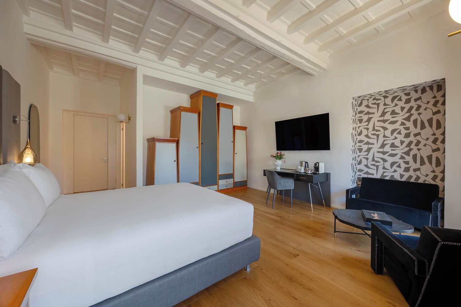 A hotel room with a large bed, a desk with a chair, a flat-screen TV, and a patterned wall.