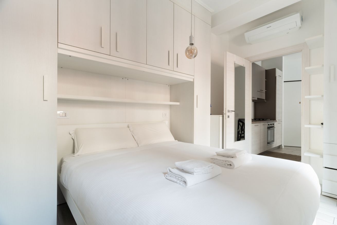A modern bedroom with a large bed, built-in wardrobe, and a kitchenette in the background.
