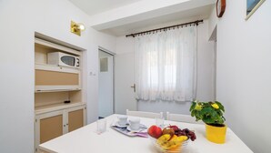 Una habitación con una mesa blanca, un frutero, una planta en maceta y un microondas en un estante.