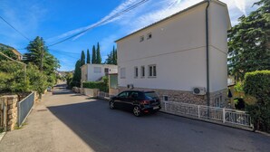 Una calle residencial con un coche negro estacionado al lado, un edificio blanco con base de piedra, y un cielo azul despejado.