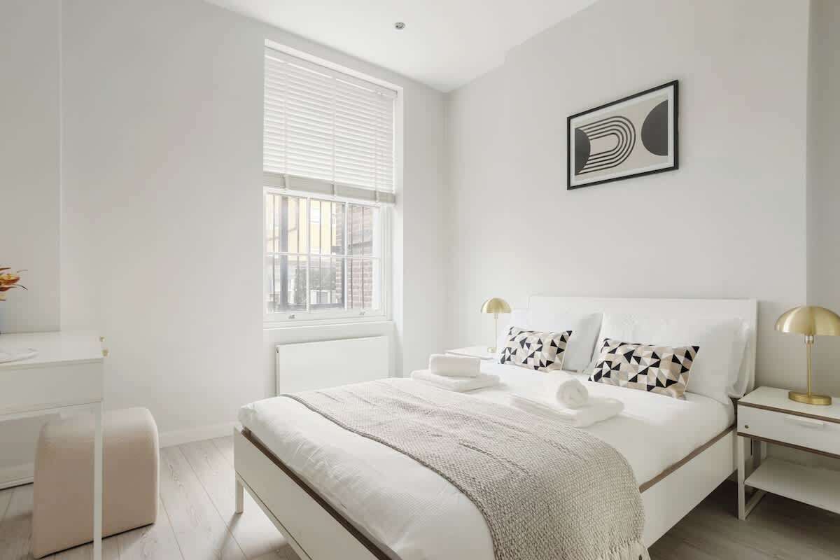 A modern bedroom with a large bed, bedside tables, a window with blinds, and a framed artwork on the wall.